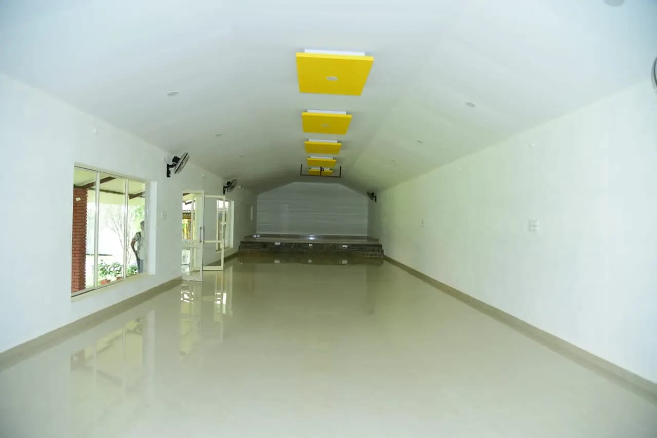 The banquet hall empty and daylit, white walls under a white false ceiling with yellow lighting drops and a low stage at the far end